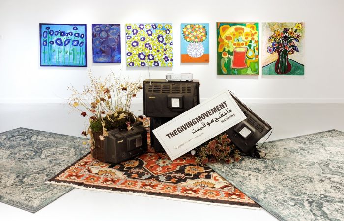 Four paintings hung up by artists from Mawaheb with three beautiful rugs along with ‘The Giving Movement’ poster in the middle - Mawaheb Art Studio for People of Determination